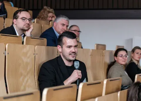 Teilnehmer stellt im Hörsaal eine Frage ins Handmikrofon, umgeben von weiteren Zuhörenden in den Sitzreihen.