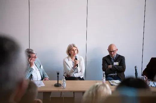 Podiumsrunde der WiföLAB-Tagung 2026: eine Rednerin mit Handmikrofon am Tisch, links und rechts zwei zuhörende Podiumsgäste.
