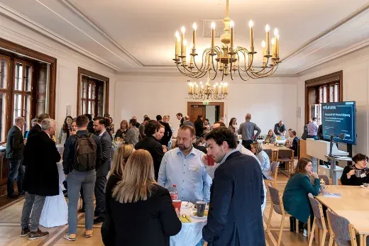 Gut gefüllter Pausensaal mit Stehtischen unter einem Kronleuchter, Teilnehmende im Gespräch.