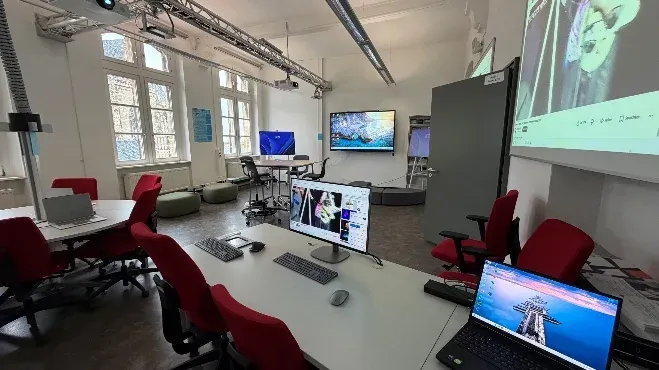 Zweiter Blick in den WiföLAB-Raum: Arbeitstische mit Bildschirmen und roten Stühlen, rechts eine bespielte Leinwand, links die hohen Fenster des Altbaus am Domplatz