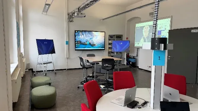 Blick in den WiföLAB-Raum: an der Decke montierte Projektoren, ein großes Wandmonitor-Display, ein mobiler Surface-Hub, im Vordergrund ein runder Tisch mit roten Stühlen und aufgeklappten Laptops